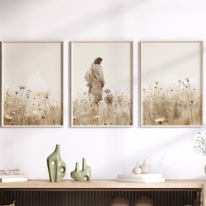 May include: A triptych of framed art prints depicting a figure with a sheep in a field, with a neutral color palette. The prints are displayed above a wooden console table with decorative vases, a basket, and a hat. The room has a neutral color scheme.