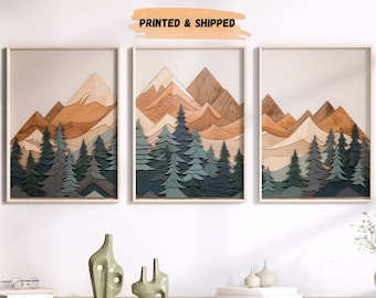 May include: Three framed prints of a layered paper art mountain scene with a woodgrain texture. The mountains are in shades of brown and orange, with green trees in the foreground. The prints are hanging on a white wall above a wooden console table.