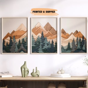 May include: Three framed prints of a layered paper art mountain scene with a woodgrain texture. The mountains are in shades of brown and orange, with green trees in the foreground. The prints are hanging on a white wall above a wooden console table.