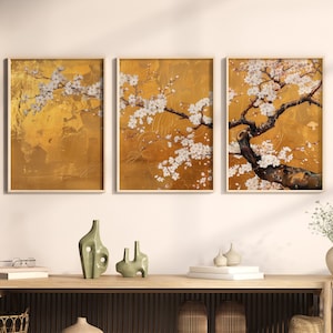 Op de afbeelding: Drie ingelijste prints van een witte kersenboom met bloesems op een gouden achtergrond. De prints hangen aan een witte muur boven een houten consoletafel met een geweven mand, twee vazen en een hoed.