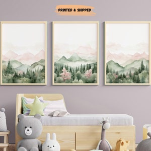 Puede incluir: Tres pinturas de acuarela enmarcadas que representan una escena de bosque con montañas rosas y verdes en el fondo. Las pinturas están colgadas en una pared de color púrpura claro sobre una cama de madera.