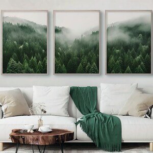 Puede incluir: Tres impresiones enmarcadas de una escena de bosque brumoso con árboles verdes. Las impresiones están colgadas en una pared blanca sobre un sofá blanco con una manta de tiro verde.