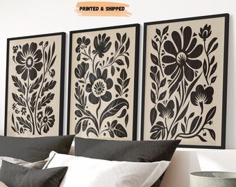 May include: Three black and white floral prints with a beige background. The prints are framed in black and are hanging on a white wall above a bed with beige linens.