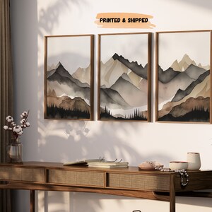 Peut inclure: Un ensemble de trois tableaux encadr&eacute;s &agrave; l'aquarelle repr&eacute;sentant une cha&icirc;ne de montagnes dans des tons de brun, gris et noir. Les tableaux sont accroch&eacute;s &agrave; un mur blanc avec une console en bois en dessous.