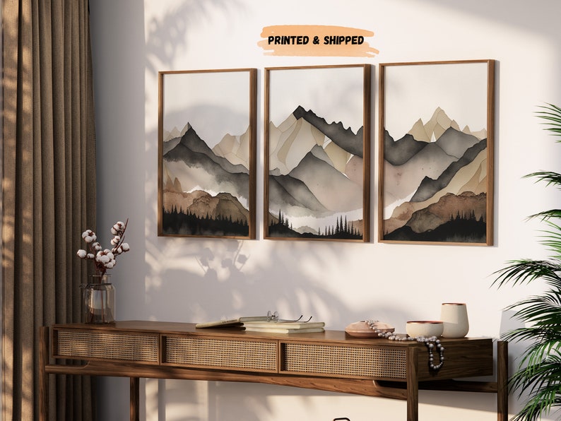 Peut inclure: Un ensemble de trois tableaux encadr&eacute;s &agrave; l'aquarelle repr&eacute;sentant une cha&icirc;ne de montagnes dans des tons de brun, gris et noir. Les tableaux sont accroch&eacute;s &agrave; un mur blanc avec une console en bois en dessous.