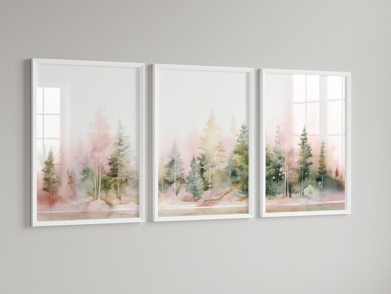 Op de afbeelding: Een set van drie ingelijste aquarellen die een bos sc&egrave;ne afbeelden in roze en groene tinten. De schilderijen zijn in witte lijsten en hangen aan een witte muur.