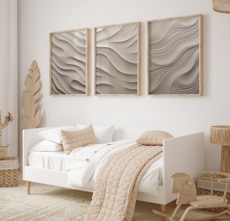 May include: A white daybed with a beige blanket and pillows, set against a white wall with three framed abstract prints of wavy lines in shades of gray.
