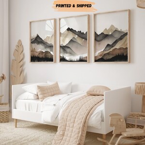 Peut inclure: Trois peintures &agrave; l'aquarelle encadr&eacute;es de cha&icirc;nes de montagnes dans des tons de brun, gris et noir. Les peintures sont accroch&eacute;es &agrave; un mur blanc au-dessus d'un lit d'appoint blanc avec une couverture beige.