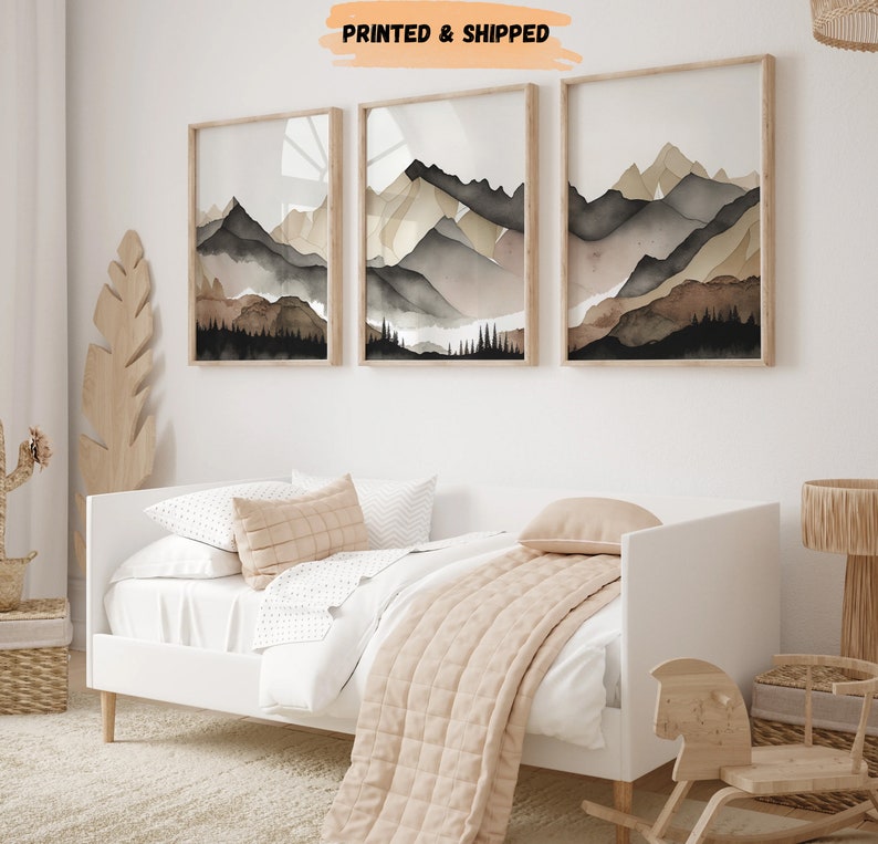 Peut inclure: Trois peintures &agrave; l'aquarelle encadr&eacute;es de cha&icirc;nes de montagnes dans des tons de brun, gris et noir. Les peintures sont accroch&eacute;es &agrave; un mur blanc au-dessus d'un lit d'appoint blanc avec une couverture beige.