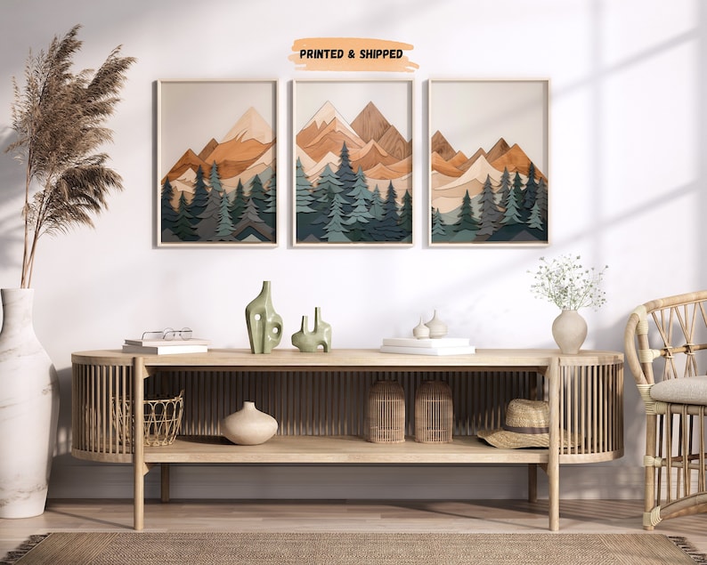 May include: Three framed prints of a layered paper art mountain scene with a woodgrain texture. The mountains are in shades of brown and orange, with green trees in the foreground. The prints are hanging on a white wall above a wooden console table.