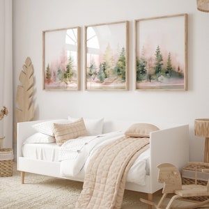 Op de afbeelding: Drie ingelijste aquarellen van een boslandschap in roze en groene tinten. De schilderijen hangen aan een witte muur boven een wit dagbed met een beige deken en kussens.