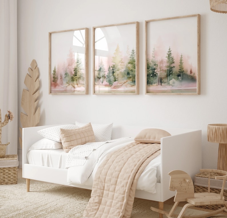 Op de afbeelding: Drie ingelijste aquarellen van een boslandschap in roze en groene tinten. De schilderijen hangen aan een witte muur boven een wit dagbed met een beige deken en kussens.