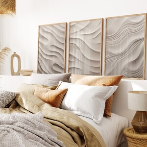 May include: A bedroom with a bed made with a brown duvet and white and brown pillows. Three framed abstract prints with a wavy pattern in shades of beige and white hang above the bed.