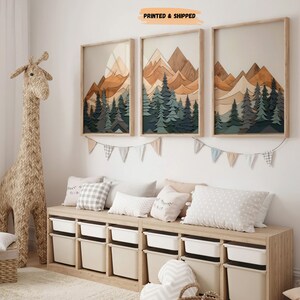 May include: Three framed wood art prints depicting a mountain range with a forest in the foreground. The prints are in shades of brown, green, and blue. The prints are hanging on a white wall above a wooden bench with white storage bins. The bench is decorated with pillows.