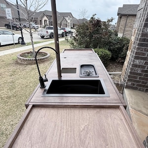 Puede incluir: Isla de cocina exterior con encimera de madera y fregadero negro. Cuenta con grifo, placa de cocina y poste met&aacute;lico. La isla est&aacute; instalada en un patio, con un barrio residencial al fondo.