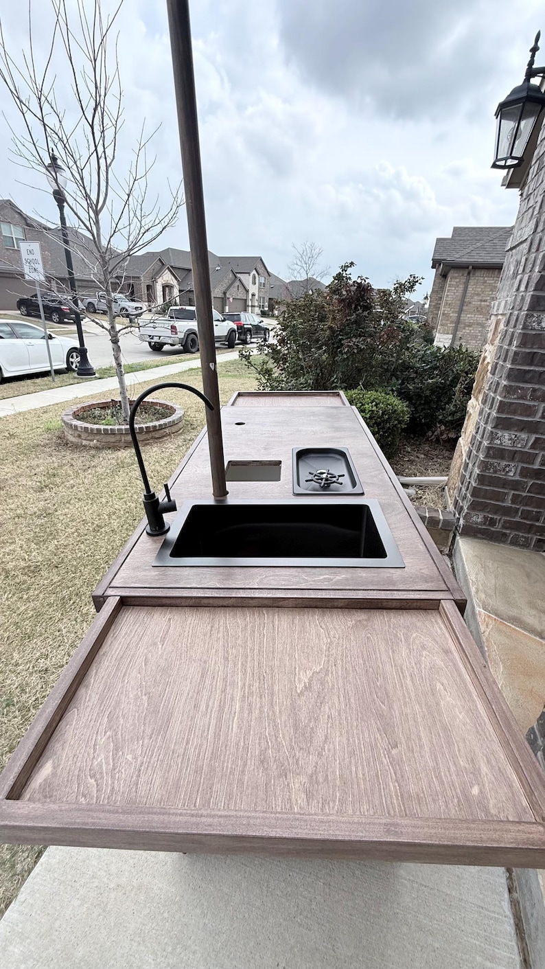 Puede incluir: Isla de cocina exterior con encimera de madera y fregadero negro. Cuenta con grifo, placa de cocina y poste met&aacute;lico. La isla est&aacute; instalada en un patio, con un barrio residencial al fondo.
