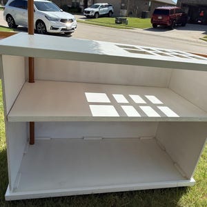 May include: A white wooden display stand with two shelves and copper-coloured support poles. The top has square cutouts. The stand is outdoors on grass.