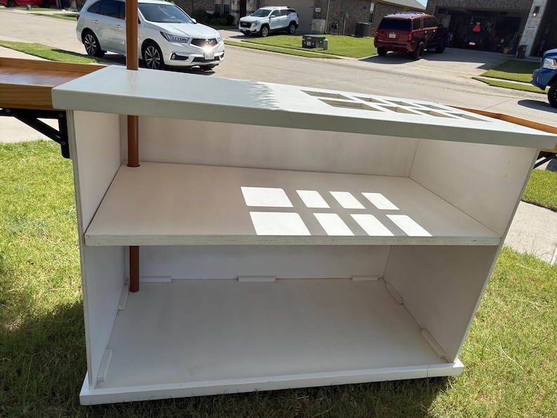 May include: A white wooden display stand with two shelves and copper-coloured support poles. The top has square cutouts. The stand is outdoors on grass.