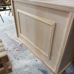 May include: An unfinished wooden cabinet, featuring a recessed rectangular panel on the front. Constructed from light-coloured wood, the cabinet has a simple design. It is likely intended for storage purposes.