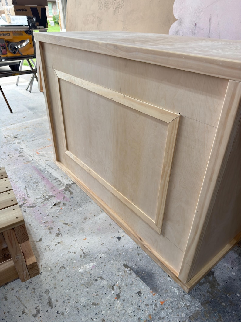 May include: An unfinished wooden cabinet, featuring a recessed rectangular panel on the front. Constructed from light-coloured wood, the cabinet has a simple design. It is likely intended for storage purposes.
