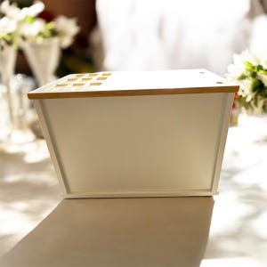 May include: A white rectangular storage box with a light wood-coloured top and accents. The box has a slightly angled design and is sitting on a table. The top has several small rectangular cutouts.