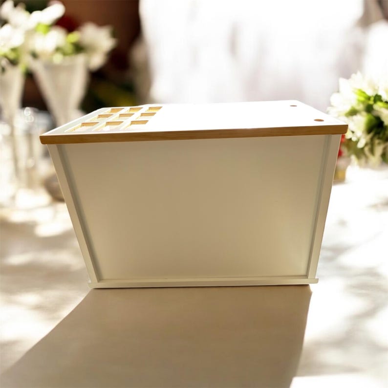 May include: A white rectangular storage box with a light wood-coloured top and accents. The box has a slightly angled design and is sitting on a table. The top has several small rectangular cutouts.