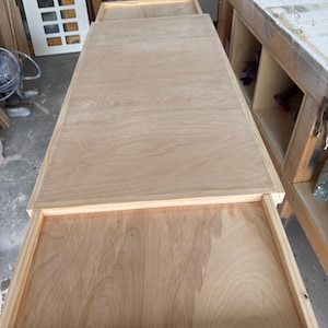 May include: A light-coloured wooden table with a drawer partially open. The table has a rectangular top and a frame around the drawer. The wood grain is visible. The table is in a workshop setting.