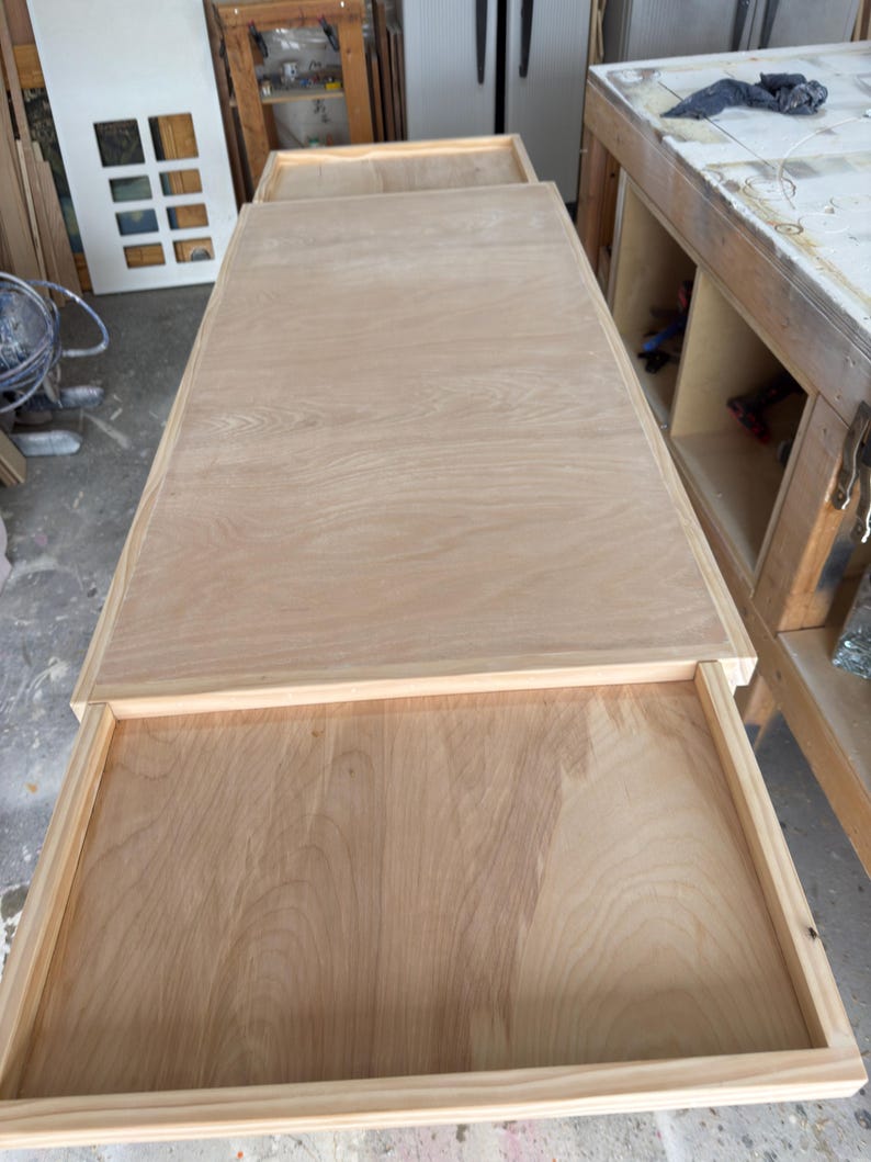 May include: A light-coloured wooden table with a drawer partially open. The table has a rectangular top and a frame around the drawer. The wood grain is visible. The table is in a workshop setting.