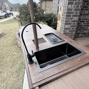 Puede incluir: Isla de cocina exterior con acabado en madera oscura. Cuenta con un fregadero negro, un grifo y una peque&ntilde;a placa de cocina. Un poste alto y oscuro se eleva desde el centro. La isla est&aacute; sobre un patio de hormig&oacute;n.
