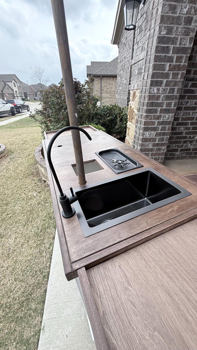 Puede incluir: Isla de cocina exterior con acabado en madera oscura. Cuenta con un fregadero negro, un grifo y una peque&ntilde;a placa de cocina. Un poste alto y oscuro se eleva desde el centro. La isla est&aacute; sobre un patio de hormig&oacute;n.