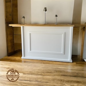 May include: A white wooden bar with a rectangular panel design. The bar is set against a light-coloured wall and a wooden shelf. The floor is made of wood. The logo "SON DOMINO WOODWORKING" is visible on the floor.