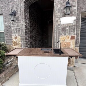 Puede incluir: Un carrito de bar exterior blanco con una encimera de madera oscura y un fregadero negro. El carrito tiene una base blanca con un dise&ntilde;o circular y est&aacute; sobre ruedas. El carrito de bar tiene dos mesas laterales plegables.