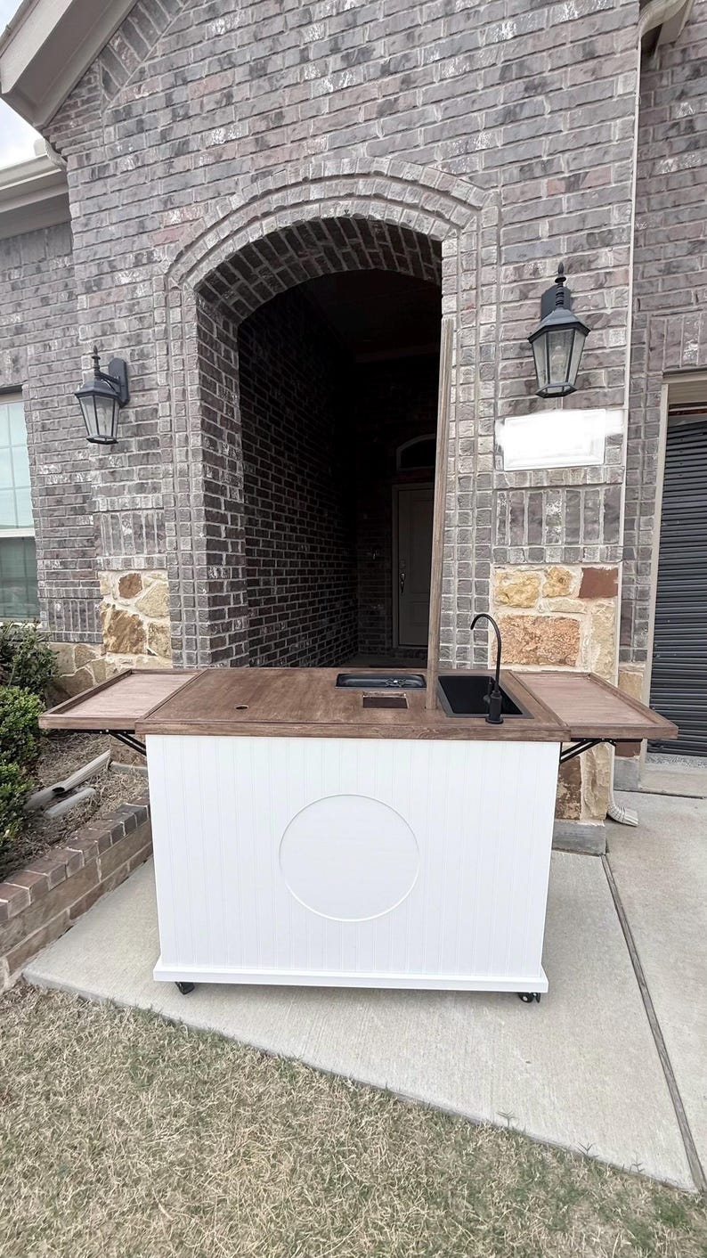 Puede incluir: Un carrito de bar exterior blanco con una encimera de madera oscura y un fregadero negro. El carrito tiene una base blanca con un dise&ntilde;o circular y est&aacute; sobre ruedas. El carrito de bar tiene dos mesas laterales plegables.