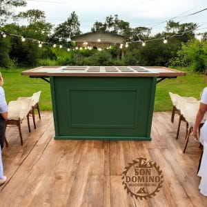 May include: A green wooden bar with a light-coloured wooden countertop. The bar has a rectangular design with a recessed panel on the front. The bar is set up outdoors on a wooden deck, with chairs and string lights in the background.