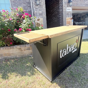Puede incluir: Una barra exterior negra con una tapa de madera clara y un estante lateral plegable. La barra presenta el texto "tabac society" en blanco. La barra est&aacute; instalada en un c&eacute;sped frente a una casa de ladrillo.
