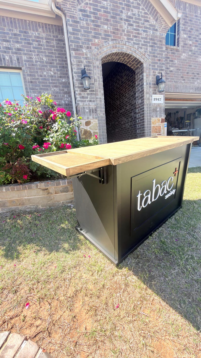 Puede incluir: Una barra exterior negra con una tapa de madera clara y un estante lateral plegable. La barra presenta el texto "tabac society" en blanco. La barra est&aacute; instalada en un c&eacute;sped frente a una casa de ladrillo.