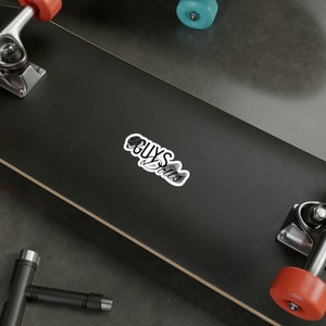 Op de afbeelding: Een zwart-witte sticker met de tekst "GUYS & Dolls" op een zwart skateboard.