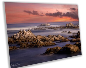 May include: A scenic photograph of a rocky coastline at sunset. The sky is a vibrant orange and pink, with clouds reflecting the warm light. The ocean is a deep blue, with waves crashing against the rocks. The rocks are a variety of browns and greys, with some covered in a light mist.