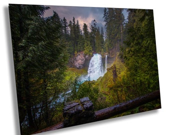 May include: A view of a waterfall cascading down a rocky cliff, surrounded by lush green trees. The waterfall is framed by a wooden railing and a stone wall.