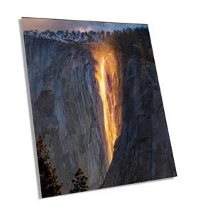 May include: A fiery orange waterfall cascades down a rocky cliff face, creating a dramatic scene in Yosemite National Park. The sun sets behind the waterfall, casting a warm glow on the surrounding landscape.
