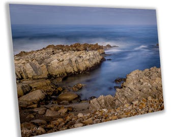 May include: A rocky coastline with a blue ocean in the background. The water is blurred, creating a sense of motion. The rocks are a mix of brown and grey, with some green algae growing on them.