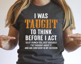 May include: A dark grey t-shirt with the text "I was taught to think before I act. So if I punch you, rest assured I've thought about it and am confident in my decision." printed in white. The text "TAUGHT" is in orange.