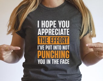 May include: Dark gray t-shirt with the text "I HOPE YOU APPRECIATE THE EFFORT I'VE PUT INTO NOT PUNCHING YOU IN THE FACE" in white and orange lettering. The shirt is being pointed at by a person.