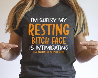 May include: Dark gray t-shirt with the text "I'M SORRY MY RESTING BITCH FACE IS INTIMIDATING I'M ACTUALLY SUPER NICE" in orange and white. The shirt is a crew neck and is being pointed at by two fingers.