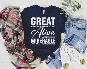 May include: A navy blue t-shirt with white text that reads "Great another day to be alive and miserable just what I always wanted". The shirt is laid out on a white surface with a plaid blanket and blue jeans.
