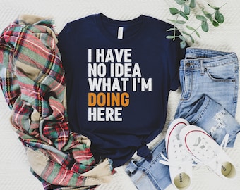 May include: A navy blue t-shirt with white text that reads "I have no idea what I'm doing here". The text is in a distressed font. The shirt is laid out on a white surface with a plaid blanket, blue jeans, and white sneakers.