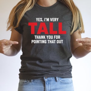 Puede incluir: Una camiseta gris oscuro con el texto "YES, I'M VERY TALL THANK YOU FOR POINTING THAT OUT" en letras blancas. La palabra "TALL" est&aacute; en rojo.
