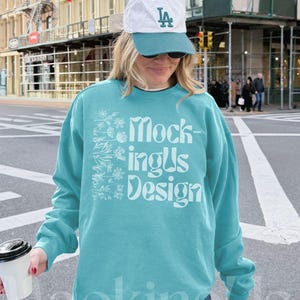 Könnte beinhalten: Ein türkisfarbenes Sweatshirt mit dem weißen Text "MockingUs Design", kombiniert mit einer weiß-türkisfarbenen Baseballkappe. Auf der Kappe stehen die Buchstaben "LA". Die Person trägt eine blaue Jeans und hält eine Kaffeetasse.