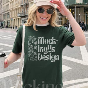 May include: Dark green t-shirt with the text "MockingUs Design" in white, paired with a beige baseball cap. The cap features the text "The Western House Casino Las Vegas Nevada". The person is wearing sunglasses and carrying a white purse.