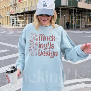 May include: A light blue sweatshirt with the text "MockingUs Design" in a reddish-brown color. The sweatshirt has a floral design on the left side. The person is wearing a white and blue baseball cap and sunglasses.